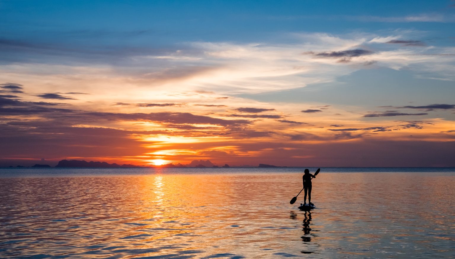 Girl on a stand up paddleboard with sunset and tropical island in the distance.